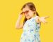 Beautiful emotional little girl isolated on yellow background. Half-lenght portrait of happy child standing and wearing a dress and red sunglasses. Concept of summer, human emotions, childhood.