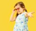 Beautiful emotional little girl isolated on yellow background. Half-lenght portrait of happy child standing and wearing a dress and red sunglasses. Concept of summer, human emotions, childhood.