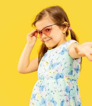 Beautiful emotional little girl isolated on yellow background. Half-lenght portrait of happy child standing and wearing a dress and red sunglasses. Concept of summer, human emotions, childhood.